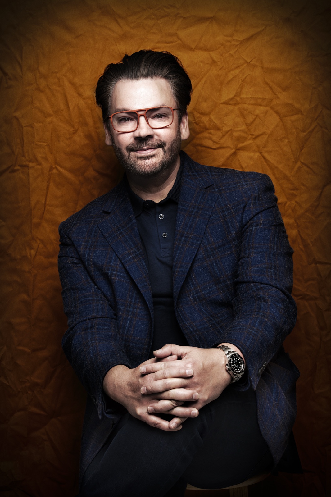 Derek seated with his hands resting together on his knees, wearing a blue jacket against a gold backdrop