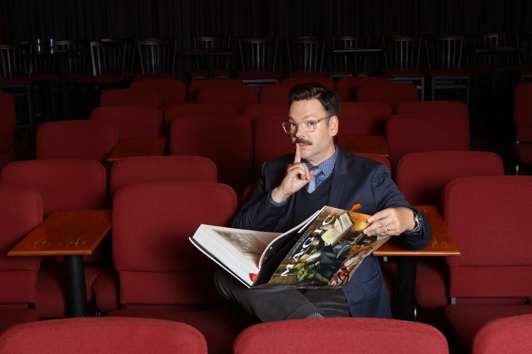 Derek seated among red theater chairs, reading a large Magic book and gesturing for quiet